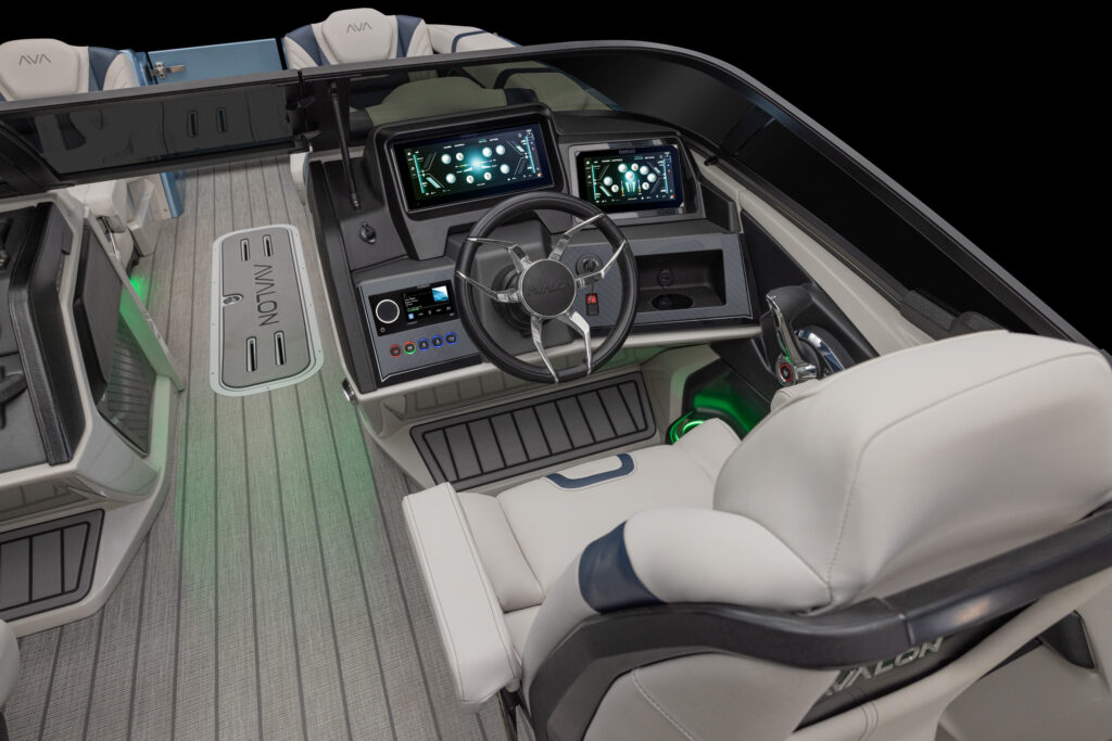 Close-up view of a modern boat's helm featuring a steering wheel, multiple digital display screens, and control panels, surrounded by plush white seating and gray flooring with ambient green lighting.