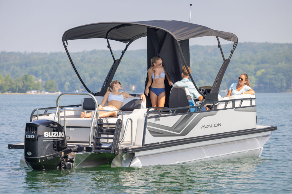 Four women relax on a pontoon boat with a black canopy, floating on calm water near a forested shoreline. Two women lounge at the back, while two sit near the helm, talking and enjoying the sunny day.