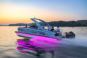 A modern pontoon boat with glowing purple LED lights speeds across a calm lake at sunset, carrying several people. Trees and the horizon are visible in the background.
