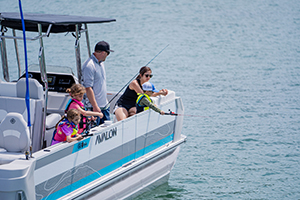 A man and three children are fishing from the side of a white and blue Avalon pontoon boat on a calm body of water. The children are wearing life jackets and appear focused on their fishing rods.