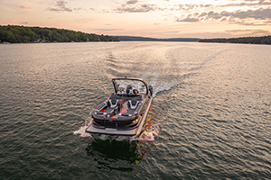 A motorboat cruises across a calm lake at sunset, leaving a wake behind. The sky is partly cloudy and the shoreline with trees is visible in the distance.