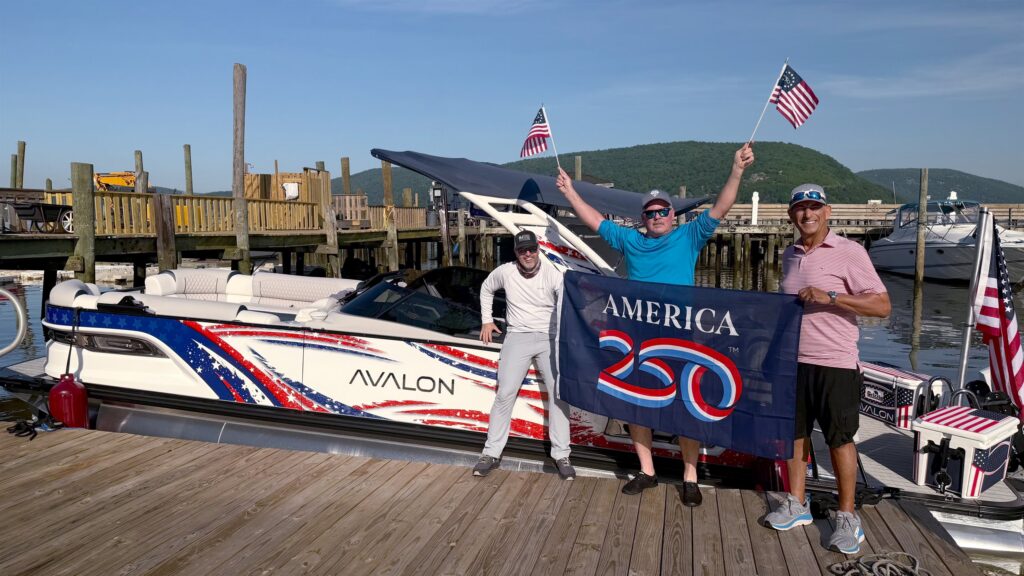 Three people pose on a dock beside the patriotic Avalon Patriot pontoon boat, holding American flags and an America 250 banner.