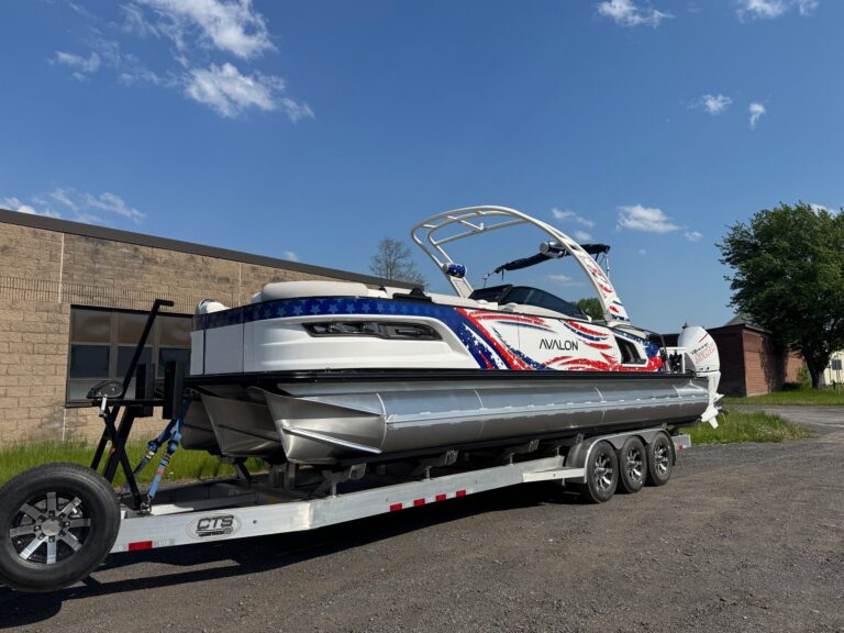 Avalon Patriot pontoon boat with patriotic red, white, and blue graphics sitting on a trailer outside a building under a blue sky.