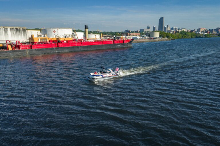 Aerial view of the Avalon Patriot pontoon boat cruising on the Hudson River near Albany, New York, with shoreline and city buildings in the distance.
